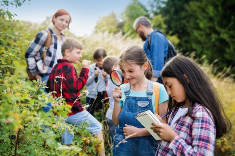 Schweizer Jugendherbergen Schulausflug Schulausflug im Grünen. Zwei Mädchen im Vordergrund stellen Untersuchungen mit Lupe und Notizblock an. Im Hintergrund sind weitere Kinder und zwei Lehrpersonen. Geschlafen wird in den Schweizer Jugendherbergen.