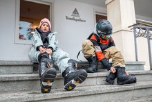 Zwei Kinder/Jugendliche sitzen vor dem Youthpalace Davos und ziehen sich Snowboard-Boots oder Skischuhe an.