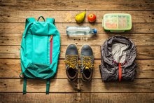 Auf einem Holzuntergrund liegen ein Rucksack, ein Paar Wanderschuhe, eine Regenjacke, eine Wasserflasche und eine Dose mit Proviant