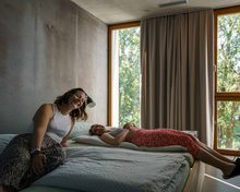 Zwei junge Frauen liegen auf einem Doppelbett in der Jugendherberge Bern. Das Zimmer ist hell, es ist Sommer und vor dem Fenster sieht man grüne Bäume.