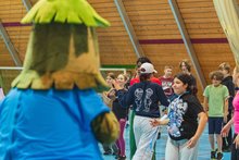 Children dance together with a Christmas tree mascot in a gymnasium. 