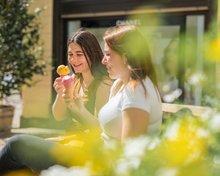 Zwei junge Frauen sitzen nebeneinander und essen Erdbeer- und Mangoeis. Im Vordergrund blühen gelbe Blumen.