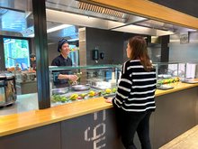 Eine junge Frau in schwarz-weiss gestreiftem Pullover steht an der Theke des Restaurant 3a in der Jugendherberge Interlaken. Ihr gegenüber ein Koch mit schwarzer Kochjacke und schwarzer Kochmütze. Er hält einen Wok am Stil und ist gerade dabei aus einer Flasche Öl in den Wok zu giessen.