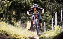 Eine junge, sportliche Frau brettert auf einem roten Mountainbike einen Berg- oder Waldweg hinunter. Sie trägt einen Helm, eine windschnittige Sonnenbrille sowie Ellbogen- und Knieschoner.