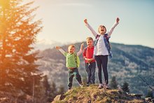 Drei Kinder, ein etwas älteres Mädchen und zwei etwas jüngere Buben, sind am Wandern, stehen auf einer kleinen Anhöhe und Jubeln vor Freude.  