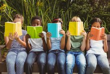 Fünf jüngere Schüler*innen sitzen auf einer Bank und lesen je ein Buch. Die Bücher halten sie so, dass ihre Gesichter verdeckt sind.