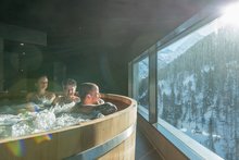 Three people, two women and one man, are sitting in the wooden barrel whirlpool. The man in the middle of the picture looks out of the large window to his right into the snow-covered mountains. The sun causes exciting lens reflections on the picture.