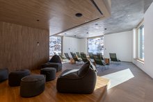 Ruhezone im Aua Grava wellnessHostel 3000 in Laax. In der Zone links sind die Wände aus Holz, auf einem Podest stehen weiche Hocker. Hinten stehen Liegestühle, sie sind in Richtung der grossen Fenster ausgerichtet. In den Fenstern sieht man das winterliche Laax.ht aus der Deckenleuchte und dem Wasser.