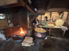 Eine junge Frau rührt, unter Anleitung des Käsermeisters, in einem Kupferkessel Milch über dem Feuer.