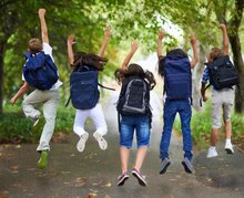 Funf Kinder oder Jugendliche sind im Wald. Sie bilden quer übers Bild eine Reihe, alle tragen sie einen Schulrucksack, hüpfen in die Luft und recken die Hände vor Freude in die Höhe.