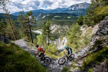 Zwei Biker fahren auf einem Biketrail. Der Trail ist eng und auf der linken Seite ist ein Abgrund. Das Foto zeigt zusätzlich die Aussicht in die Rheinschlucht in Graubünden. Das Foto wurde auf dem Alpine Circle aufgenommen.