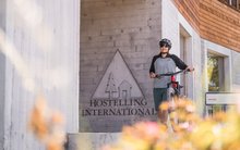 Eine junge Bikerin steht mit ihrem Bike vor dem Eingangsbereich der Jugendherberge Gtsaad Saanenland. Das Hostelling International Logo ist im Hintergrund zu erkennen. 