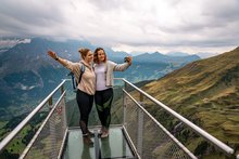 Zwei junge Frauen stehen auf dem First Cliff Walk und machen ein Selfie. Im Hintergrund das Bergpanorama.